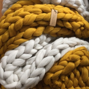 May include: A chunky knit blanket in white and mustard yellow. The blanket is made of thick, soft yarn, with a tag that reads "WOOLLY CLOUD 100% MERINO WOOL". The blanket is folded, showing off the braided texture.