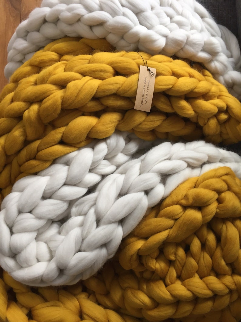 May include: A chunky knit blanket in white and mustard yellow. The blanket is made of thick, soft yarn, with a tag that reads "WOOLLY CLOUD 100% MERINO WOOL". The blanket is folded, showing off the braided texture.