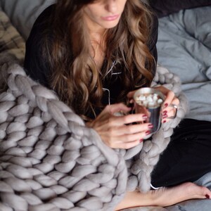 May include: A person wrapped in a large, chunky knit gray blanket, holding a mug of hot chocolate with marshmallows. The blanket is draped over a bed with gray bedding. The person is wearing black pajamas.