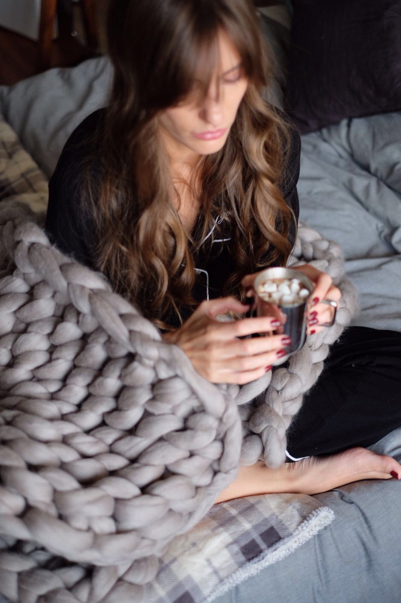 May include: A person wrapped in a large, chunky knit gray blanket, holding a mug of hot chocolate with marshmallows. The blanket is draped over a bed with gray bedding. The person is wearing black pajamas.