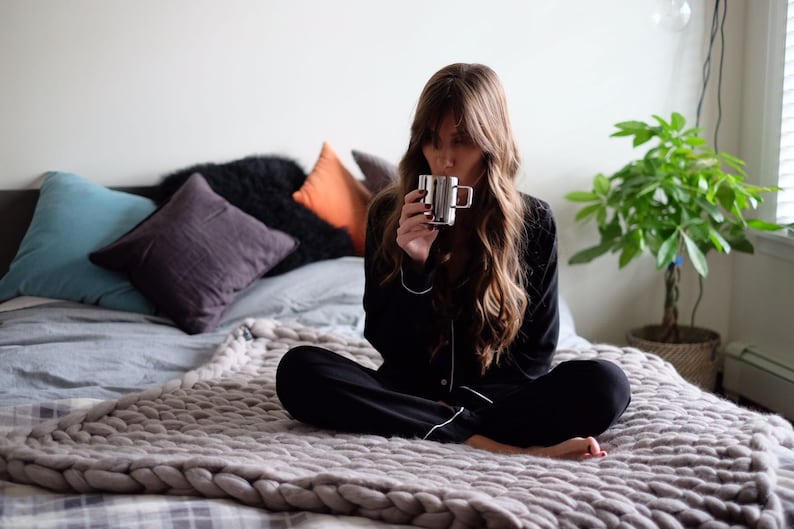 May include: A person in black pajamas with white trim sits on a bed, holding a silver mug. The bed is covered with a gray chunky knit blanket and pillows in various colors. A potted plant is in the background.