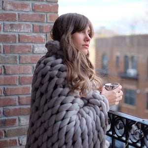 May include: A person wearing a chunky, gray knit blanket wrap, holding a silver mug on a balcony. The background features a brick wall and a view of buildings. The blanket appears to be made of thick wool.