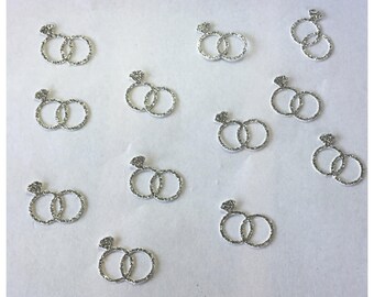 May include: A collection of eleven silver-colored glittery embellishments, each shaped like two interlocking rings with a small diamond-like accent at the top. These flat, decorative pieces are arranged in a scattered pattern across the frame.