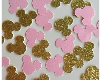 May include: A collection of scattered confetti pieces shaped like mouse heads. The confetti consists of a mix of solid light pink cardstock cutouts and gold glitter-covered cutouts, all featuring the iconic three-circle silhouette design.