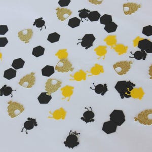 May include: A collection of paper confetti pieces in the shapes of black hexagons, yellow bees, black bees, and gold glitter beehives scattered across a surface. The assorted shapes are flat and vary in color, texture, and design elements.