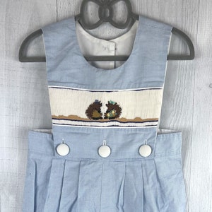 May include: Light blue and white striped romper with a smocked panel featuring embroidered hedgehogs. The romper has white buttons and a pleated design. The garment is hanging on a gray hanger.