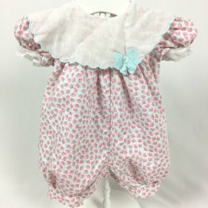 Puede incluir: Un pelele de bebé vintage con cuello festoneado blanco y lazo azul claro. El pelele es blanco con un estampado de fresas rosas y mangas abullonadas. Tiene un bajo abombado y cierres de botón.