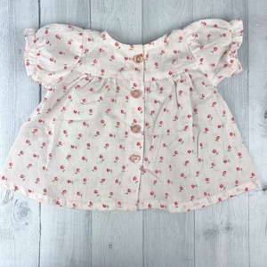 May include: A light pink baby top with short, ruffled sleeves and a button-up front. The top is covered in small red floral patterns and has matching pink buttons. The fabric appears to be lightweight cotton.
