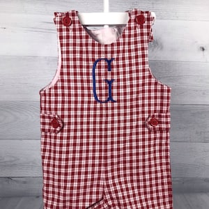 Puede incluir: Romper a cuadros rojos y blancos con una letra "G" bordada en azul. El romper tiene botones rojos en los hombros y los lados. El forro interior es de tela rosa claro. La prenda está colgada de una percha blanca.