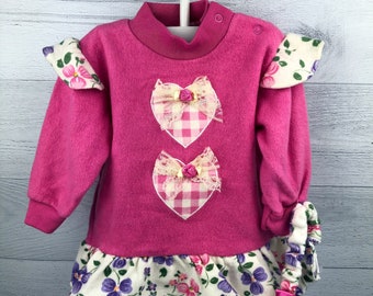 May include: A vibrant pink fleece dress with a high neck and long sleeves. The dress has two heart-shaped appliqués with gingham and lace, and a floral print ruffle. The sleeves have floral cuffs and a pink bow.