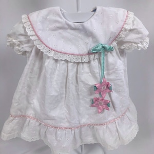 May include: White eyelet dress with a ruffled collar and hem, trimmed with pink ribbon. The dress features short sleeves and is adorned with a mint green bow and pink flower accents. The dress is hanging on a white hanger.