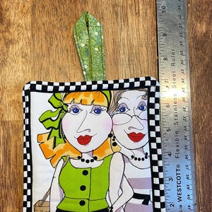 May include: A black and white checkered fabric pot holder with a green, white, and orange fabric applique of two women with exaggerated features. The women are wearing green and gray dresses and have black hair.