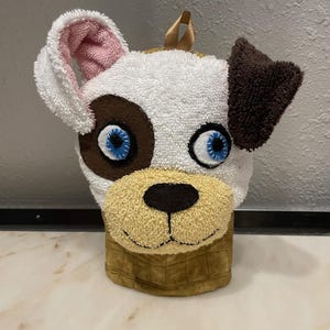 May include: A white and brown dog-shaped oven mitt with pink and brown ears and blue eyes. The mitt is made of a soft, plush material and has a brown and tan checkered pattern on the bottom.