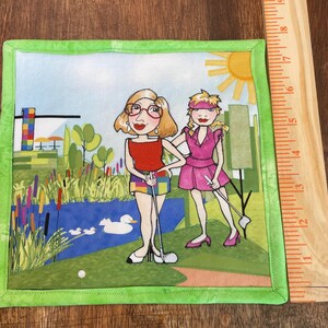 May include: Square fabric art with a green border featuring two women playing golf. The women are depicted in a cartoon style, standing on a green golf course with a pond, ducks, and a sun in the background. The artwork is approximately 8 inches square.