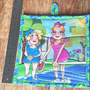 May include: Square quilted potholder featuring a colorful illustration of two women playing pickleball. The design includes a blue and green border, a hanging loop, and a whimsical depiction of a pickleball court.