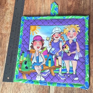 May include: A quilted pot holder featuring a colorful illustration of three women playing pickleball. The design includes a purple background with a green and blue border, a bench, and accessories. The pot holder has a loop for hanging.