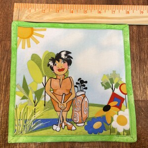 May include: Square fabric art featuring a cartoon golfer in an orange outfit, holding a golf club. The background depicts a golf course with trees, flowers, and a golf bag. The artwork is framed with a green border.