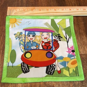 May include: Square fabric coaster with a whimsical golf theme. The coaster features a cartoon illustration of two women in a golf cart, surrounded by flowers and trees. The border is a bright green.