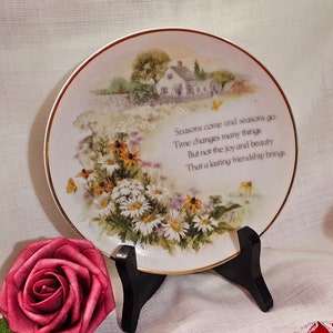 Peut inclure: Une assiette en porcelaine blanche avec un bord doré présente un motif floral coloré et une scène de cottage. L'assiette porte une citation qui dit : "Les saisons vont et viennent Le temps change beaucoup de choses Mais pas la joie et la beauté Que l'amitié durable apporte"