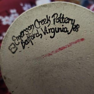 Op de afbeelding: Close-up van de onderkant van een keramisch stuk met de tekst "Emerson Creek Pottery Bedford, Virginia". Het aardewerk is lichtbeige met zwarte handgeschreven tekst en een rode streep.