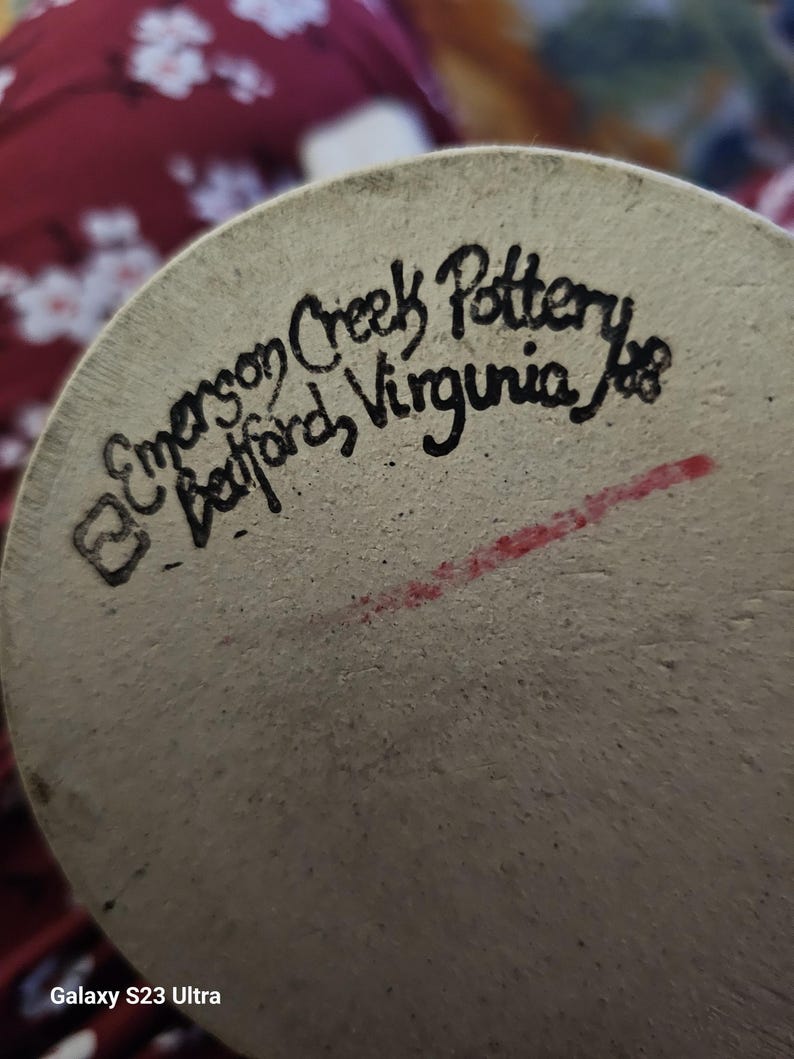 Op de afbeelding: Close-up van de onderkant van een keramisch stuk met de tekst "Emerson Creek Pottery Bedford, Virginia". Het aardewerk is lichtbeige met zwarte handgeschreven tekst en een rode streep.