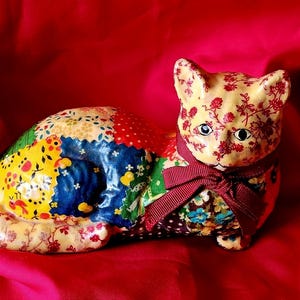 May include: A ceramic cat figurine with a patchwork design. The cat's head is cream-colored with red floral patterns, and it wears a burgundy bow. The body features a colorful patchwork of floral and patterned fabrics, set against a vibrant red background.