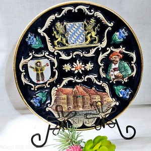May include: A black and gold ceramic plate with a decorative design featuring a building, a coat of arms, and a flower. The plate is on a black metal stand. The text "MUNCHEN" is visible on the plate.