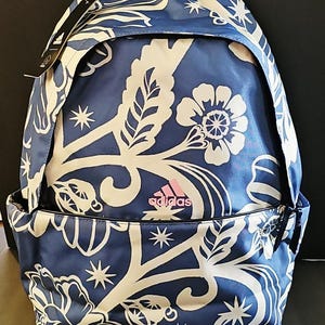 Puede incluir: Mochila azul con un estampado floral blanco y el logotipo de Adidas en rosa. La mochila tiene un asa superior, correas ajustables y un bolsillo frontal. El diseño incluye flores, hojas y elementos en forma de estrella.