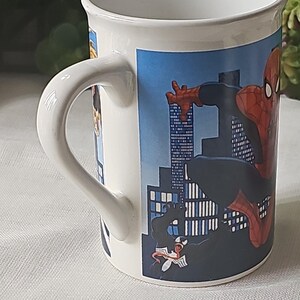 Puede incluir: Taza de cer&aacute;mica blanca con una imagen colorida de Spiderman y Venom frente a un horizonte urbano. La taza tiene un asa negra.