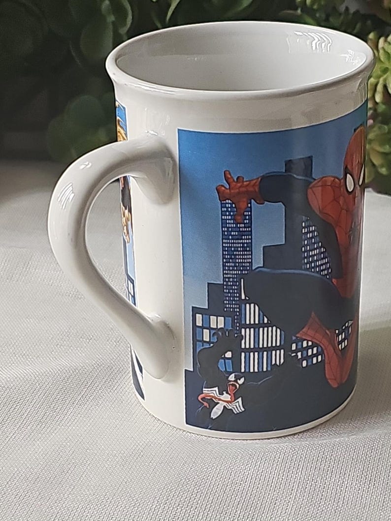 Puede incluir: Taza de cer&aacute;mica blanca con una imagen colorida de Spiderman y Venom frente a un horizonte urbano. La taza tiene un asa negra.