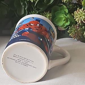 Puede incluir: Una taza de cer&aacute;mica blanca con un dise&ntilde;o azul y rojo con Spiderman. La taza tiene un asa y dice "MARVEL" en la parte inferior. La parte inferior de la taza tambi&eacute;n incluye el texto "TM & &copy; 2016 Marvel & Subs. &copy;2016 Frankford Candy LLC Made in China MICROWAVE AND DISHWASHER SAFE MFG-Z 20160620".