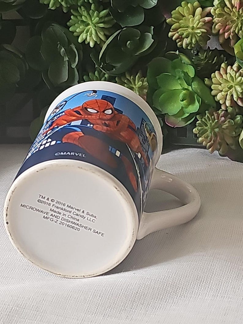 Puede incluir: Una taza de cer&aacute;mica blanca con un dise&ntilde;o azul y rojo con Spiderman. La taza tiene un asa y dice "MARVEL" en la parte inferior. La parte inferior de la taza tambi&eacute;n incluye el texto "TM & &copy; 2016 Marvel & Subs. &copy;2016 Frankford Candy LLC Made in China MICROWAVE AND DISHWASHER SAFE MFG-Z 20160620".