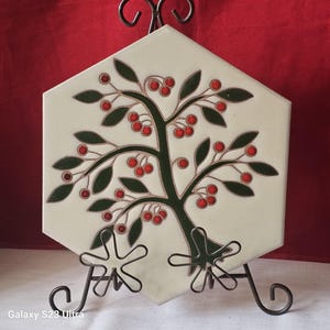 Puede incluir: Azulejo cerámico hexagonal decorativo con diseño de árbol, con hojas verdes y bayas rojas. El azulejo se exhibe sobre un soporte de hierro forjado. El fondo es de color rojo liso.
