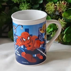 Puede incluir: Una taza de cer&aacute;mica blanca con un dise&ntilde;o de Spiderman azul, rojo y negro. La taza tiene un asa y el logotipo de Marvel.