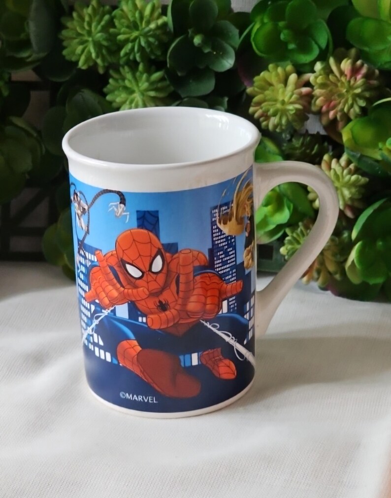 Puede incluir: Una taza de cer&aacute;mica blanca con un dise&ntilde;o de Spiderman azul, rojo y negro. La taza tiene un asa y el logotipo de Marvel.