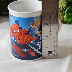 Puede incluir: Una taza de cer&aacute;mica blanca con una ilustraci&oacute;n azul, roja y amarilla de Spiderman balance&aacute;ndose por una ciudad. La taza tiene el texto "OMARVEL" en la parte inferior.