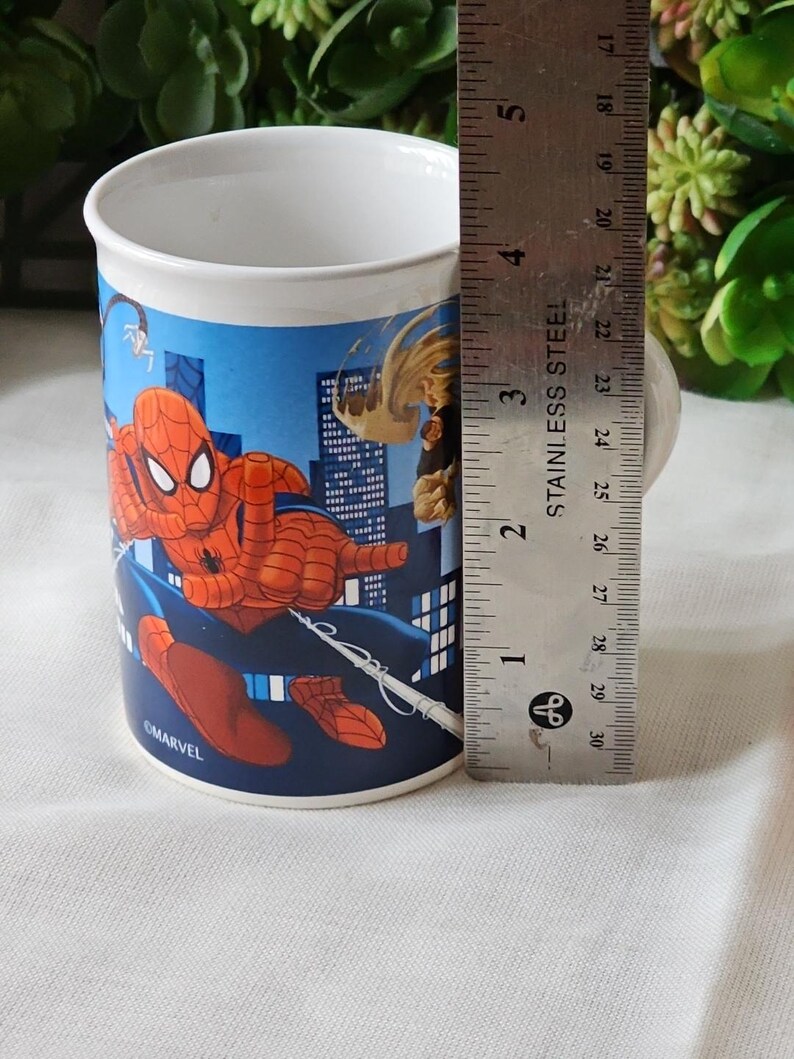 Puede incluir: Una taza de cer&aacute;mica blanca con una ilustraci&oacute;n azul, roja y amarilla de Spiderman balance&aacute;ndose por una ciudad. La taza tiene el texto "OMARVEL" en la parte inferior.