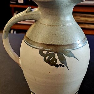 Op de afbeelding: Handgemaakte keramische kan in lichtgroen en cr&egrave;me. Het heeft een gebogen handvat en een tuit. De voorkant is versierd met een donkergroen bloemmotief. Deze kan is een decoratief item.
