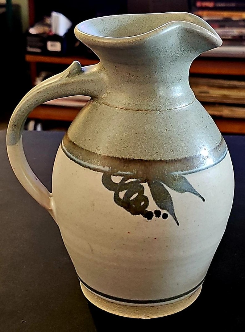 Op de afbeelding: Handgemaakte keramische kan in lichtgroen en cr&egrave;me. Het heeft een gebogen handvat en een tuit. De voorkant is versierd met een donkergroen bloemmotief. Deze kan is een decoratief item.