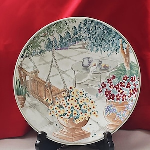 May include: A decorative plate depicting a patio scene with a swing, table, and potted flowers. The plate has a white background with a painted design in green, brown, yellow, and red. The plate is displayed on a black stand.