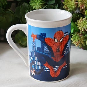 Puede incluir: Taza de cer&aacute;mica blanca con una imagen colorida de Spiderman balance&aacute;ndose en un horizonte de ciudad. Venom est&aacute; en el fondo.
