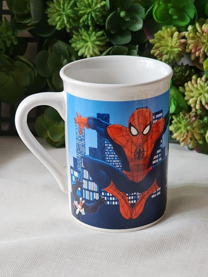 Puede incluir: Taza de cer&aacute;mica blanca con una imagen colorida de Spiderman balance&aacute;ndose en un horizonte de ciudad. Venom est&aacute; en el fondo.