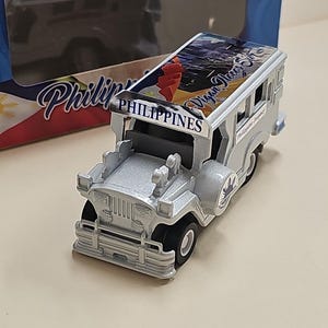 Puede incluir: Un jeep de juguete plateado con un diseño azul y blanco en el techo. El jeep tiene las palabras "Vigan Flores" y "Filipinas" impresas en el lateral.