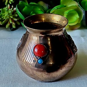 May include: A small, round, brass pot with a decorative design. The pot has three inlaid stones: two blue and one red.