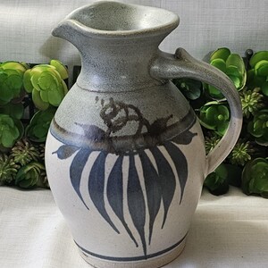Op de afbeelding: Handgemaakte keramische kan met een grijze en witte glazuur. De kan heeft een tuit en een handvat en is versierd met blauwe bladvormige patronen. Het ontwerp doet denken aan een rustieke boerderijstijl.