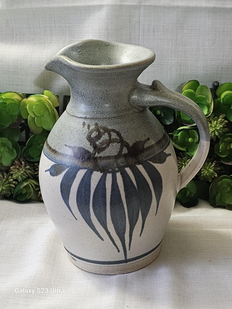 Op de afbeelding: Handgemaakte keramische kan met een grijze en witte glazuur. De kan heeft een tuit en een handvat en is versierd met blauwe bladvormige patronen. Het ontwerp doet denken aan een rustieke boerderijstijl.
