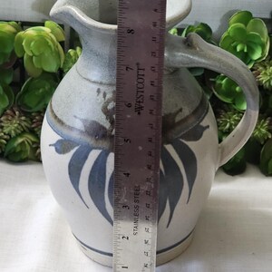 Op de afbeelding: Een keramische kan met een handvat, tuit en bladeren. De kan is voornamelijk wit met blauwe en grijze accenten. De hoogte van de kan is ongeveer 20 cm.