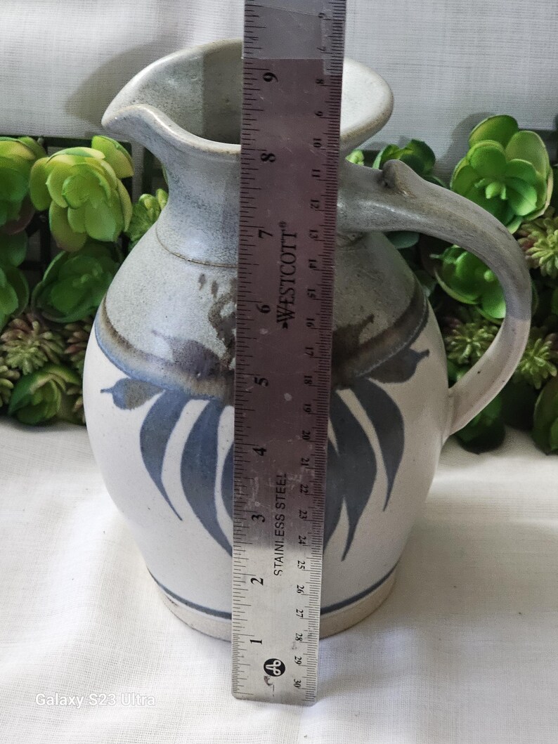 Op de afbeelding: Een keramische kan met een handvat, tuit en bladeren. De kan is voornamelijk wit met blauwe en grijze accenten. De hoogte van de kan is ongeveer 20 cm.