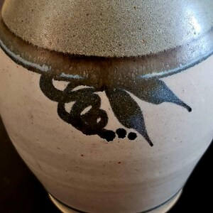 Op de afbeelding: Een keramische vaas met een afgeronde body en een smalle hals. De vaas heeft een lichtbeige basis met een donkerbruine band en een lichtgroene bovenkant. Een zwart bloemmotief siert de zijkant van de vaas. De vaas is waarschijnlijk handgemaakt.