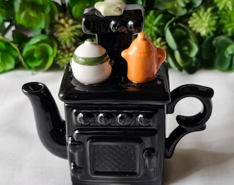 Może przedstawiać: Czarny ceramiczny czajnik w kształcie starej kuchenki z białym i zielonym czajnikiem oraz pomarańczowym czajnikiem na górze.